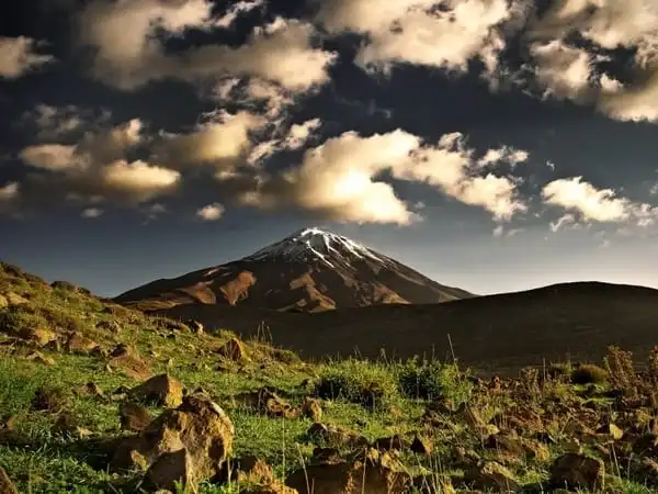 A scenic view of Mount Kilimanjaro, its snow-capped peak rising above a rolling, grassy foreground with scattered rocks, under a dramatic sky filled with white and gray clouds.
